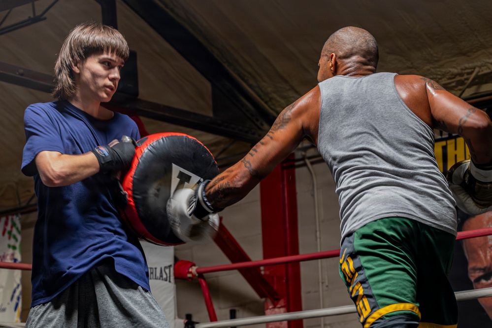 Two individuals are training in a boxing gym inside a boxing ring. A younger person wearing a blue t-shirt holds a large punch mitt while an older man in a gray tank top and green shorts throws a punch into it. The setting appears to be a well-equipped boxing gym with Everlast branding visible in the background.