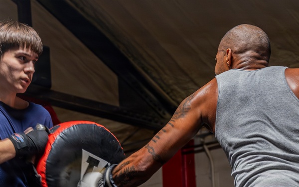 Two individuals are training in a boxing gym inside a boxing ring. A younger person wearing a blue t-shirt holds a large punch mitt while an older man in a gray tank top and green shorts throws a punch into it. The setting appears to be a well-equipped boxing gym with Everlast branding visible in the background.