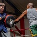 Two individuals are training in a boxing gym inside a boxing ring. A younger person wearing a blue t-shirt holds a large punch mitt while an older man in a gray tank top and green shorts throws a punch into it. The setting appears to be a well-equipped boxing gym with Everlast branding visible in the background.