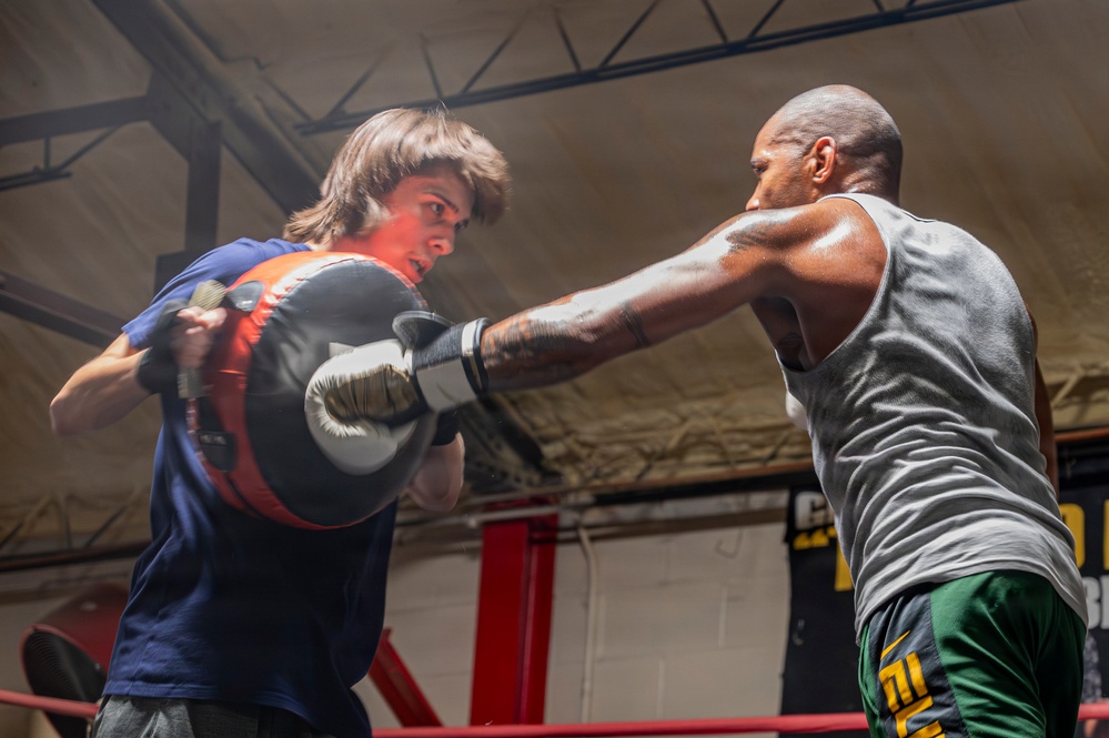 Two individuals are sparring in a boxing ring inside what appears to be a gym or training facility. One person wearing a blue t-shirt and boxing gloves is blocking a punch from a muscular opponent in a gray tank top and green shorts. The action is captured mid-punch, conveying intensity and motion.
