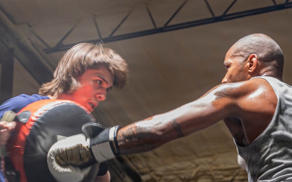 Two individuals are sparring in a boxing ring inside what appears to be a gym or training facility. One person wearing a blue t-shirt and boxing gloves is blocking a punch from a muscular opponent in a gray tank top and green shorts. The action is captured mid-punch, conveying intensity and motion.