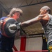 Two individuals are sparring in a boxing ring inside what appears to be a gym or training facility. One person wearing a blue t-shirt and boxing gloves is blocking a punch from a muscular opponent in a gray tank top and green shorts. The action is captured mid-punch, conveying intensity and motion.
