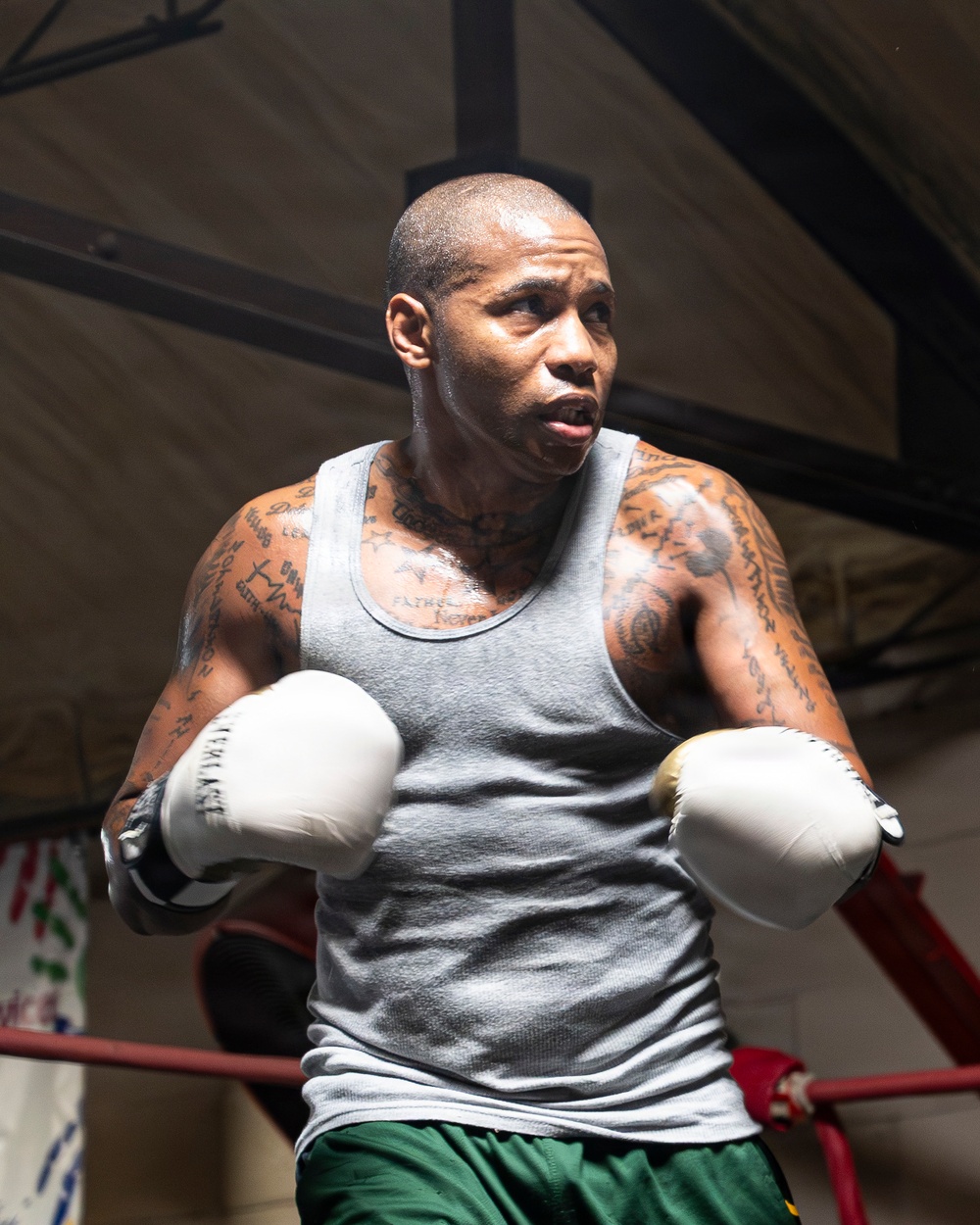 A muscular male boxer is training inside a boxing ring, wearing white boxing gloves and a gray sleeveless shirt with green shorts. He is in a fighting stance with both gloves raised, appearing focused and intense. His arms and neck are heavily tattooed, and he shows visible signs of physical exertion.