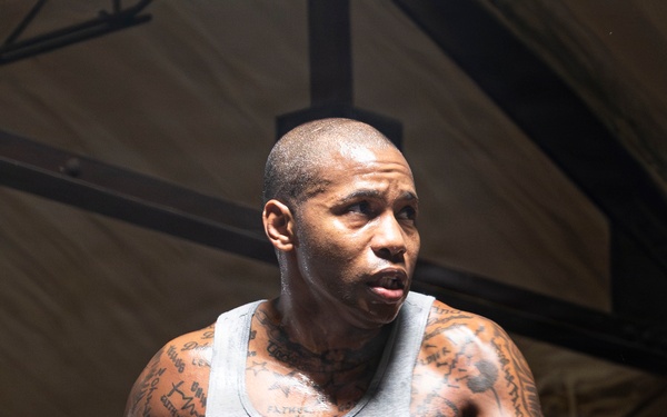 A muscular male boxer is training inside a boxing ring, wearing white boxing gloves and a gray sleeveless shirt with green shorts. He is in a fighting stance with both gloves raised, appearing focused and intense. His arms and neck are heavily tattooed, and he shows visible signs of physical exertion.