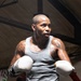 A muscular male boxer is training inside a boxing ring, wearing white boxing gloves and a gray sleeveless shirt with green shorts. He is in a fighting stance with both gloves raised, appearing focused and intense. His arms and neck are heavily tattooed, and he shows visible signs of physical exertion.