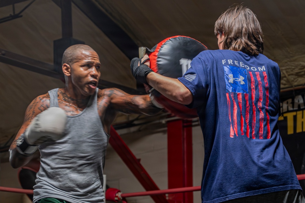 A boxer wearing a gray tank top and white boxing gloves is training in a boxing ring, throwing a punch at focus mitts held by a trainer. The trainer, facing away from the camera, is wearing a navy blue Under Armour 'Freedom' t-shirt featuring an American flag design. The image captures an active training session in what appears to be a boxing gym.