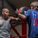 A boxer wearing a gray tank top and white boxing gloves is training in a boxing ring, throwing a punch at focus mitts held by a trainer. The trainer, facing away from the camera, is wearing a navy blue Under Armour 'Freedom' t-shirt featuring an American flag design. The image captures an active training session in what appears to be a boxing gym.