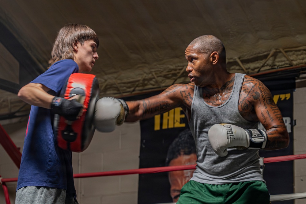 Two individuals are training in a boxing gym inside a boxing ring. A heavily tattooed man wearing a gray tank top and green shorts is throwing a punch with Everlast boxing gloves at a red focus mitt held by a younger person in a blue t-shirt. A boxing-related poster is visible on the wall in the background.