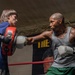 Two individuals are training in a boxing gym inside a boxing ring. A heavily tattooed man wearing a gray tank top and green shorts is throwing a punch with Everlast boxing gloves at a red focus mitt held by a younger person in a blue t-shirt. A boxing-related poster is visible on the wall in the background.