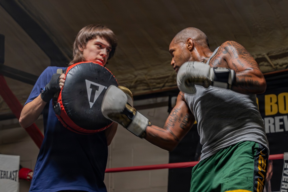Two individuals are training in a boxing gym inside a ring with red ropes. One person wearing a blue shirt holds a Title-brand punch mitt while the other, a heavily tattooed man in a gray tank top and green shorts, throws a punch. The setting appears to be an indoor boxing gym with Everlast branding visible in the background.