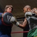 Two individuals are training in a boxing gym inside a ring with red ropes. One person wearing a blue shirt holds a Title-brand punch mitt while the other, a heavily tattooed man in a gray tank top and green shorts, throws a punch. The setting appears to be an indoor boxing gym with Everlast branding visible in the background.