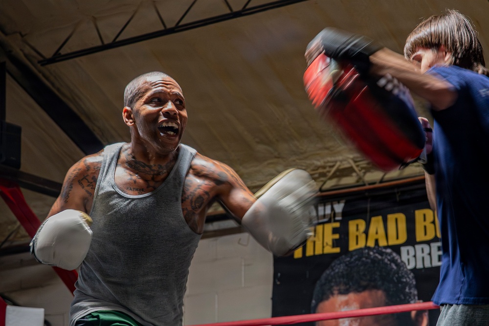 Two individuals are sparring or training in a boxing gym. The primary subject, a heavily tattooed muscular man wearing a gray tank top and white boxing gloves, is throwing a punch with an intense expression. A poster on the wall in the background partially reads 'THE BAD BO...' alongside a face portrait.