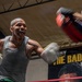 Two individuals are sparring or training in a boxing gym. The primary subject, a heavily tattooed muscular man wearing a gray tank top and white boxing gloves, is throwing a punch with an intense expression. A poster on the wall in the background partially reads 'THE BAD BO...' alongside a face portrait.