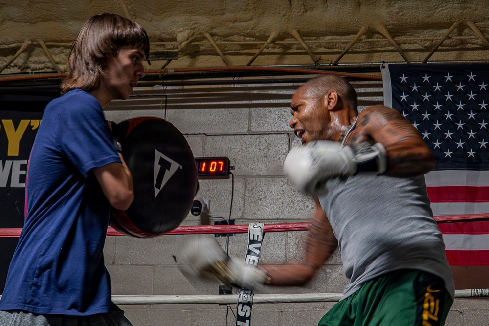 Two individuals are training in a boxing gym, with one person holding a Title brand punch mitt while the other throws punches wearing white boxing gloves. The boxer on the right, wearing a gray shirt and green shorts, is captured mid-punch with motion blur indicating fast movement. An American flag and a digital round timer reading '1:07' are visible on the cinder block wall behind them.
