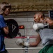 Two individuals are training in a boxing gym, with one person holding a Title brand punch mitt while the other throws punches wearing white boxing gloves. The boxer on the right, wearing a gray shirt and green shorts, is captured mid-punch with motion blur indicating fast movement. An American flag and a digital round timer reading '1:07' are visible on the cinder block wall behind them.