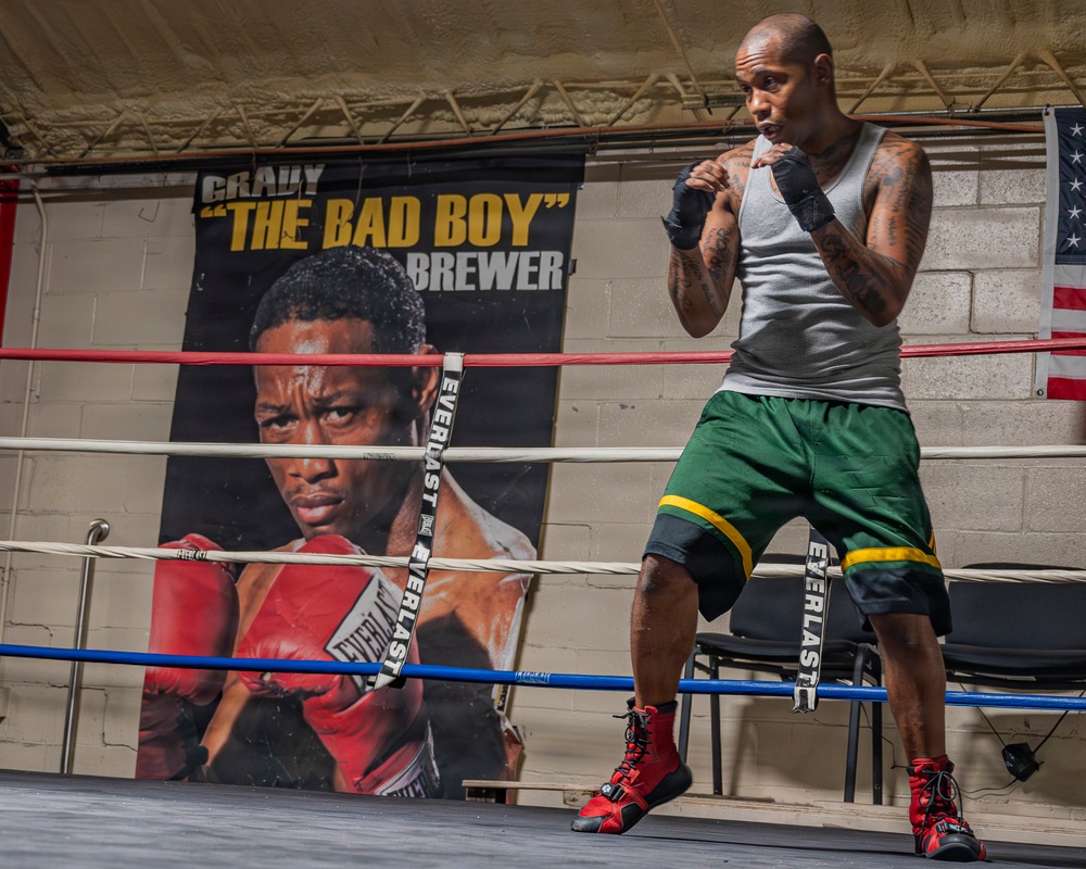 A tattooed male boxer is training in a boxing ring, shown in a fighting stance with hand wraps on both fists. Behind him is a large promotional banner reading 'Grady "The Bad Boy" Brewer' featuring a close-up portrait of a boxer wearing Everlast gloves. The setting appears to be an indoor boxing gym with an American flag visible on the wall.