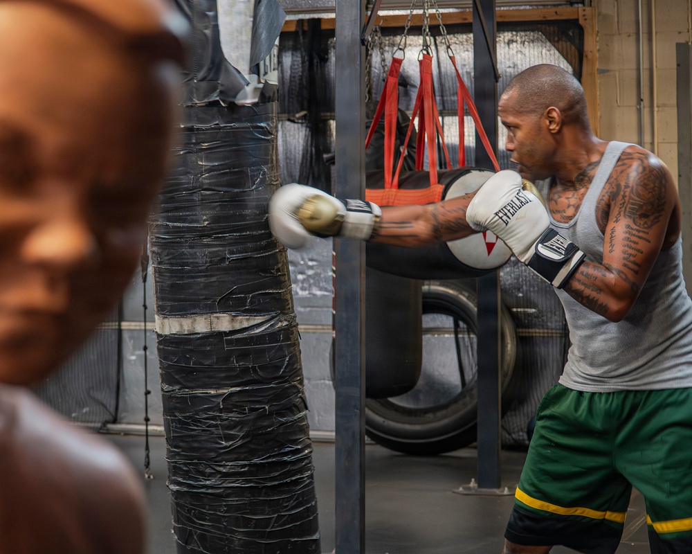 A heavily tattooed male boxer is training in a gym, striking a heavily taped heavy bag with white Everlast boxing gloves. The gym environment appears worn and industrial, featuring multiple hanging bags, old tires, and a metal support structure. A blurred figure or mannequin is visible in the foreground left.