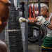 A heavily tattooed male boxer is training in a gym, striking a heavily taped heavy bag with white Everlast boxing gloves. The gym environment appears worn and industrial, featuring multiple hanging bags, old tires, and a metal support structure. A blurred figure or mannequin is visible in the foreground left.