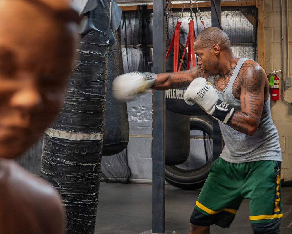 A heavily tattooed male boxer is training at a boxing gym, throwing a punch at a heavily worn and taped heavy bag. He is wearing white Everlast boxing gloves, a gray tank top, and green shorts with yellow stripes. The gym environment shows multiple heavy bags, chains, red straps, and a fire extinguisher mounted on the wall.