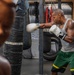 A heavily tattooed male boxer is training at a boxing gym, throwing a punch at a heavily worn and taped heavy bag. He is wearing white Everlast boxing gloves, a gray tank top, and green shorts with yellow stripes. The gym environment shows multiple heavy bags, chains, red straps, and a fire extinguisher mounted on the wall.