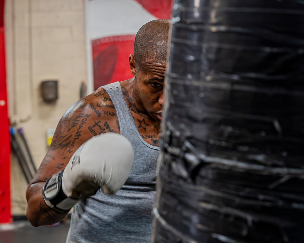 A heavily tattooed male boxer is training on a large black heavy bag in what appears to be a boxing gym. He is wearing white boxing gloves and a gray sleeveless shirt, captured mid-punch with visible motion blur on his glove. The image is taken from a close angle with the heavy bag partially obscuring his face.