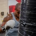 A heavily tattooed male boxer is training on a large black heavy bag in what appears to be a boxing gym. He is wearing white boxing gloves and a gray sleeveless shirt, captured mid-punch with visible motion blur on his glove. The image is taken from a close angle with the heavy bag partially obscuring his face.