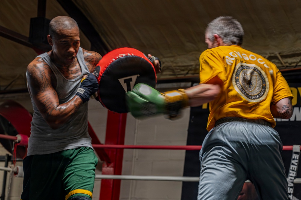 Two men are training in a boxing gym inside a boxing ring. A tattooed boxer wearing a gray tank top and green shorts is throwing a punch at a red focus mitt held by a trainer in a yellow 'Mongoose' shirt and gray pants. The motion blur on the punch indicates high-speed impact during a pad work session.