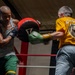 Two men are training in a boxing gym inside a boxing ring. A tattooed boxer wearing a gray tank top and green shorts is throwing a punch at a red focus mitt held by a trainer in a yellow 'Mongoose' shirt and gray pants. The motion blur on the punch indicates high-speed impact during a pad work session.