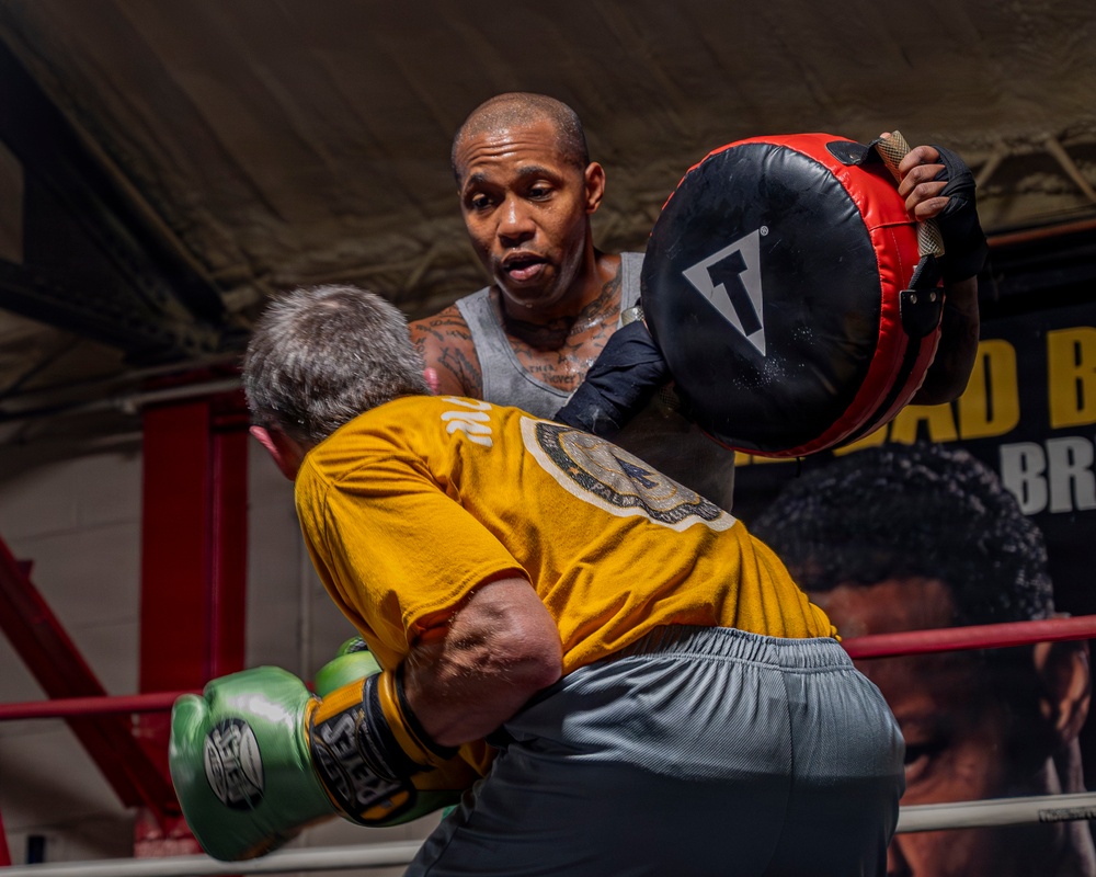 Two individuals are training in a boxing ring. One person wearing a gray tank top and visible tattoos holds a circular punch mitt (branded 'Title'), while a second person in a yellow shirt and green boxing gloves throws a punch. The setting appears to be an indoor boxing gym with red ring ropes and promotional banners visible in the background.