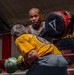 Two individuals are training in a boxing ring. One person wearing a gray tank top and visible tattoos holds a circular punch mitt (branded 'Title'), while a second person in a yellow shirt and green boxing gloves throws a punch. The setting appears to be an indoor boxing gym with red ring ropes and promotional banners visible in the background.