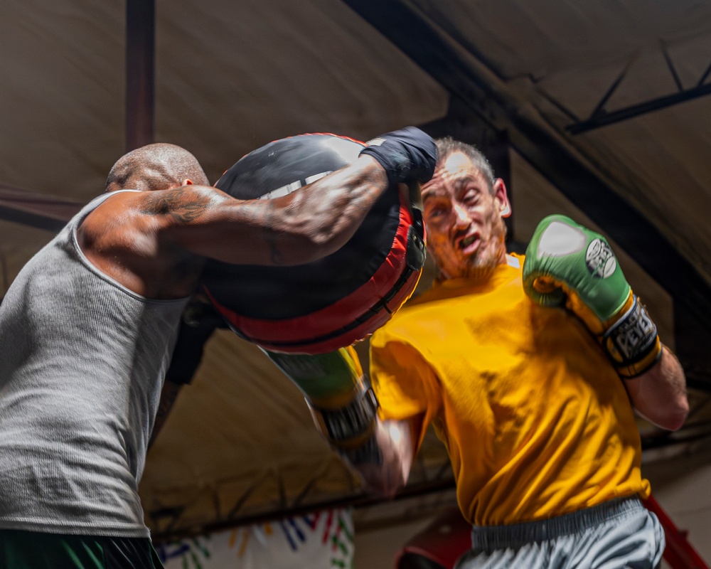 Two boxers are engaged in an intense sparring session inside what appears to be a boxing gym or training facility. The boxer on the left, wearing a gray tank top, is delivering a powerful punch with a large padded mitt or bag, making contact with the face/head area of the boxer on the right. The boxer on the right, wearing a yellow shirt and green boxing gloves, is absorbing or reacting to the impact.