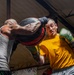 Two boxers are engaged in an intense sparring session inside what appears to be a boxing gym or training facility. The boxer on the left, wearing a gray tank top, is delivering a powerful punch with a large padded mitt or bag, making contact with the face/head area of the boxer on the right. The boxer on the right, wearing a yellow shirt and green boxing gloves, is absorbing or reacting to the impact.