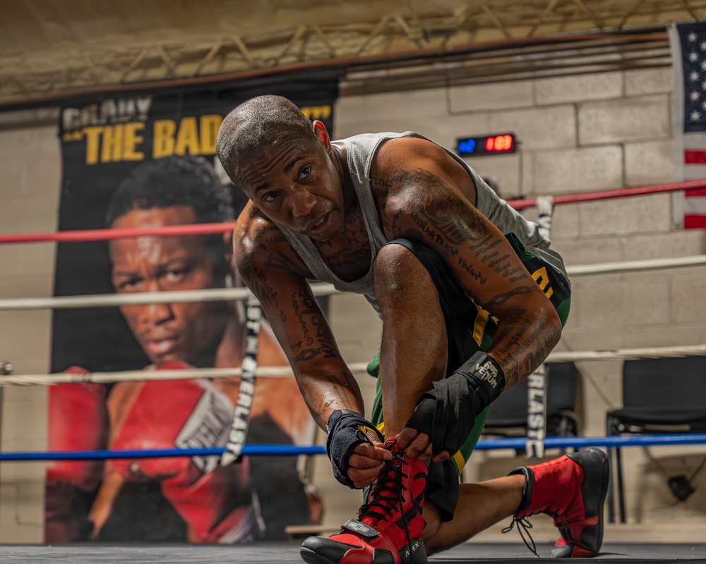 A heavily tattooed male boxer is kneeling in a boxing ring, tying the laces on his red and black boxing shoes. He is wearing a gray sleeveless shirt, green and yellow shorts, and has hand wraps on his wrists. The setting is a boxing gym with a promotional banner, an American flag, a digital timer, and Everlast-branded ring ropes visible in the background.