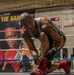 A heavily tattooed male boxer is kneeling in a boxing ring, tying the laces on his red and black boxing shoes. He is wearing a gray sleeveless shirt, green and yellow shorts, and has hand wraps on his wrists. The setting is a boxing gym with a promotional banner, an American flag, a digital timer, and Everlast-branded ring ropes visible in the background.