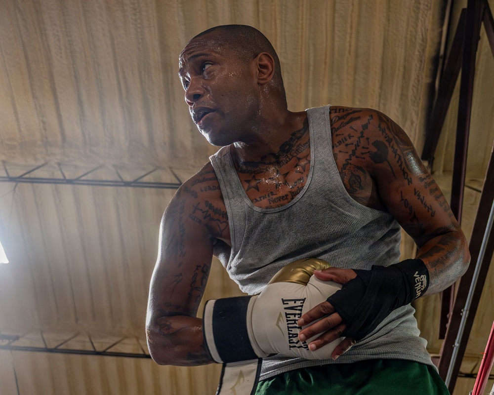 A heavily tattooed male boxer is photographed from a low angle inside what appears to be a boxing gym or ring. He is visibly sweating, suggesting intense physical exertion, and is wearing Everlast boxing gloves along with Venum hand wraps. His expression is focused and intense as he looks off to one side.