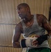 A heavily tattooed male boxer is photographed from a low angle inside what appears to be a boxing gym or ring. He is visibly sweating, suggesting intense physical exertion, and is wearing Everlast boxing gloves along with Venum hand wraps. His expression is focused and intense as he looks off to one side.