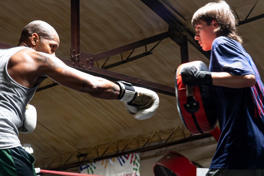 Two individuals are engaged in a boxing training session inside what appears to be a gym or boxing facility. An adult male on the left is throwing a punch with white boxing gloves, while a younger male on the right holds a red and black punch mitt to receive the strike. The setting suggests a coaching or mentoring dynamic between the two participants.