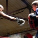 Two individuals are engaged in a boxing training session inside what appears to be a gym or boxing facility. An adult male on the left is throwing a punch with white boxing gloves, while a younger male on the right holds a red and black punch mitt to receive the strike. The setting suggests a coaching or mentoring dynamic between the two participants.