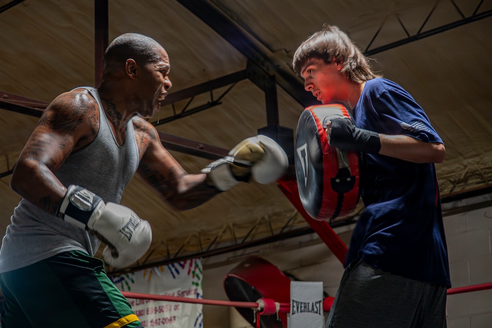 Two individuals are training in a boxing gym, with one person throwing punches wearing Everlast boxing gloves while the other holds a focus mitt/punch pad. The older man on the left has visible tattoos and is wearing a gray tank top and green shorts, while the younger person on the right is wearing a blue t-shirt and holding a training pad. The setting appears to be an indoor boxing gym with a ring, red ropes, and an Everlast banner visible in the background.