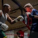 Two individuals are training in a boxing gym, with one person throwing punches wearing Everlast boxing gloves while the other holds a focus mitt/punch pad. The older man on the left has visible tattoos and is wearing a gray tank top and green shorts, while the younger person on the right is wearing a blue t-shirt and holding a training pad. The setting appears to be an indoor boxing gym with a ring, red ropes, and an Everlast banner visible in the background.