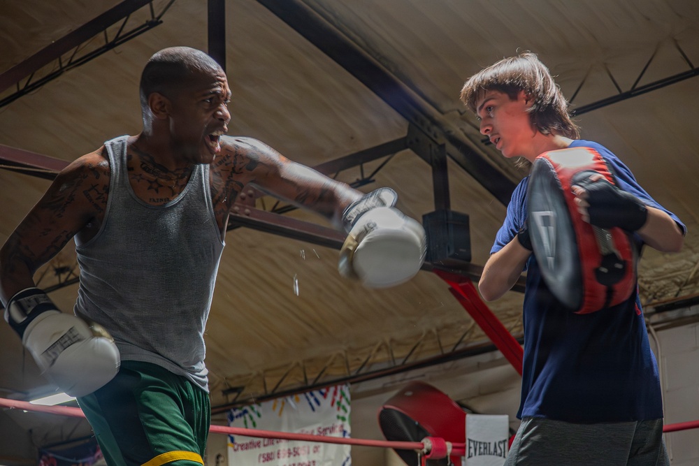 Two individuals are training in a boxing gym inside a boxing ring. The man on the left, wearing a gray tank top and green shorts with extensive tattoos, is throwing a punch while wearing white boxing gloves. The younger person on the right is holding red and black focus mitts to receive the punch, suggesting a trainer-athlete or sparring dynamic.