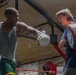 Two individuals are training in a boxing gym inside a boxing ring. The man on the left, wearing a gray tank top and green shorts with extensive tattoos, is throwing a punch while wearing white boxing gloves. The younger person on the right is holding red and black focus mitts to receive the punch, suggesting a trainer-athlete or sparring dynamic.