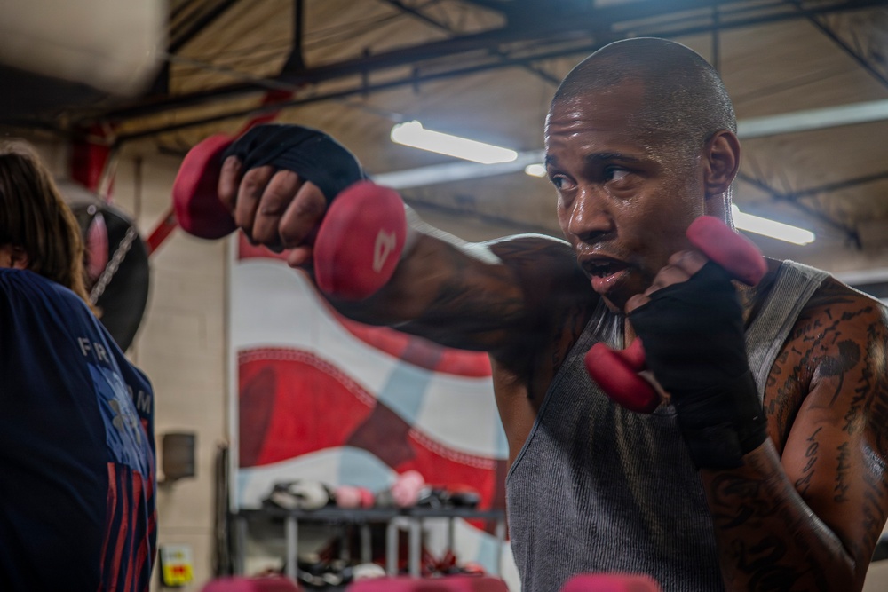 A muscular man with tattoos on his arms is training in a boxing gym, shadowboxing or drilling with small red dumbbells while wearing hand wraps. He is dressed in a gray sleeveless shirt and appears to be sweating, indicating intense physical exertion. Another person is partially visible to the left, and the gym environment includes fluorescent lighting, a mural, and boxing equipment in the background.