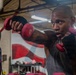 A muscular man with tattoos on his arms is training in a boxing gym, shadowboxing or drilling with small red dumbbells while wearing hand wraps. He is dressed in a gray sleeveless shirt and appears to be sweating, indicating intense physical exertion. Another person is partially visible to the left, and the gym environment includes fluorescent lighting, a mural, and boxing equipment in the background.