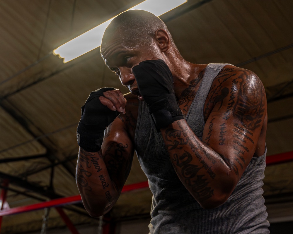 A heavily tattooed male boxer is photographed in a fighting stance inside what appears to be a boxing gym or training facility. He is wearing black hand wraps and a gray sleeveless shirt, with his fists raised in a defensive guard position. The image is dramatically lit from above, highlighting sweat on his skin and creating strong contrast.