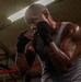 A heavily tattooed male boxer is photographed in a fighting stance inside what appears to be a boxing gym or training facility. He is wearing black hand wraps and a gray sleeveless shirt, with his fists raised in a defensive guard position. The image is dramatically lit from above, highlighting sweat on his skin and creating strong contrast.