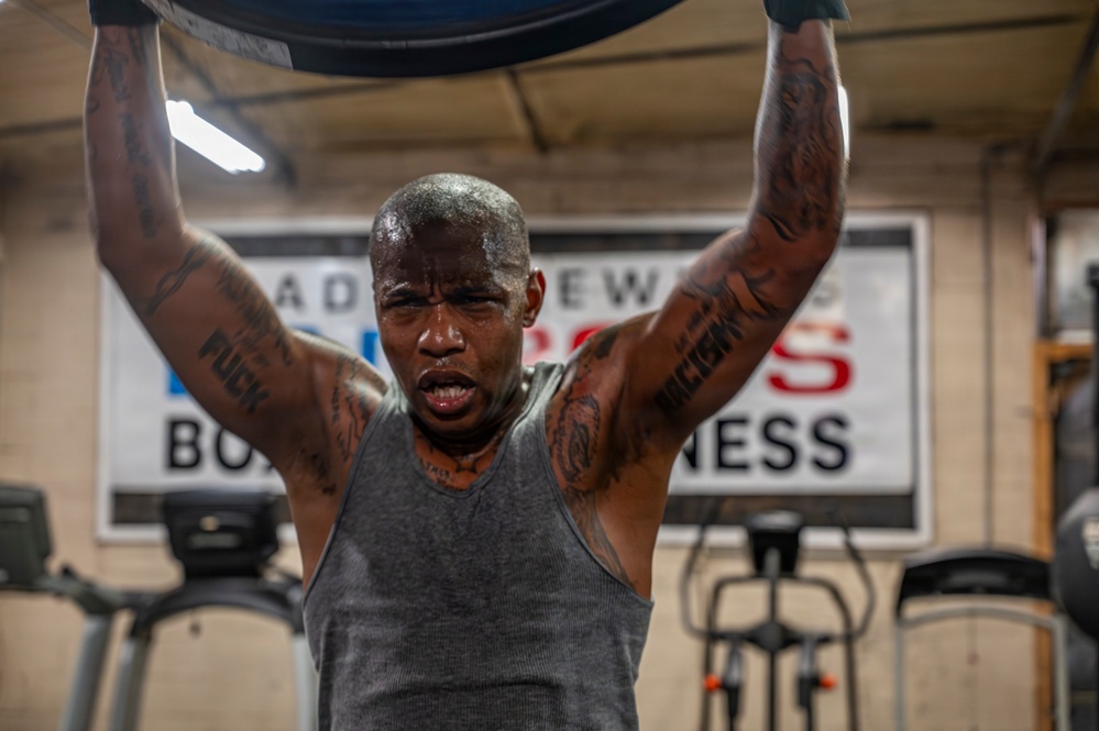 A heavily tattooed man is working out intensely in a boxing/fitness gym, lifting a large pad or equipment overhead with both hands raised. He is visibly sweating and exerting significant effort, wearing a gray sleeveless tank top. The background shows gym equipment including treadmills and a partially visible sign reading 'BOX[ING] FITNESS'.