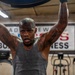 A heavily tattooed man is working out intensely in a boxing/fitness gym, lifting a large pad or equipment overhead with both hands raised. He is visibly sweating and exerting significant effort, wearing a gray sleeveless tank top. The background shows gym equipment including treadmills and a partially visible sign reading 'BOX[ING] FITNESS'.