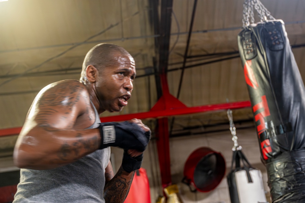 A muscular, bald male boxer is training intensely at a boxing gym, working a heavy bag with hand wraps on his fists. He is shirtless except for a gray tank top and has visible tattoos on his arms and torso. The image captures motion blur on his hands, suggesting powerful, fast movement during the workout.
