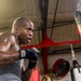 A muscular, bald male boxer is training intensely at a boxing gym, working a heavy bag with hand wraps on his fists. He is shirtless except for a gray tank top and has visible tattoos on his arms and torso. The image captures motion blur on his hands, suggesting powerful, fast movement during the workout.